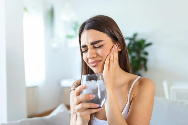 A woman drinking a cold glass of water and wincing in pain while holding her cheek.