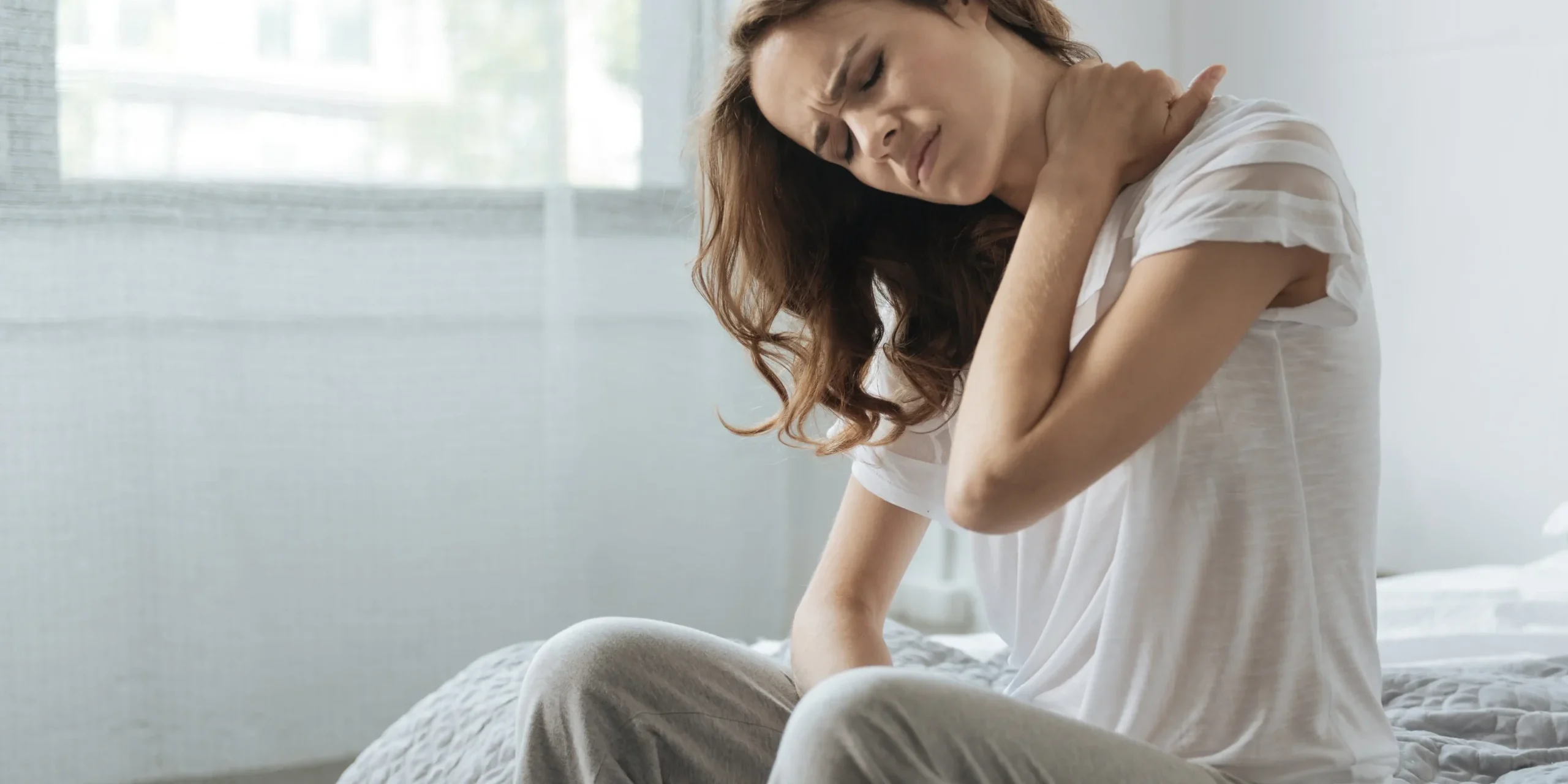 Woman seated on bed holding her neck wincing in pain.