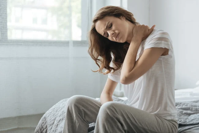 Woman seated on bed holding her neck wincing in pain.