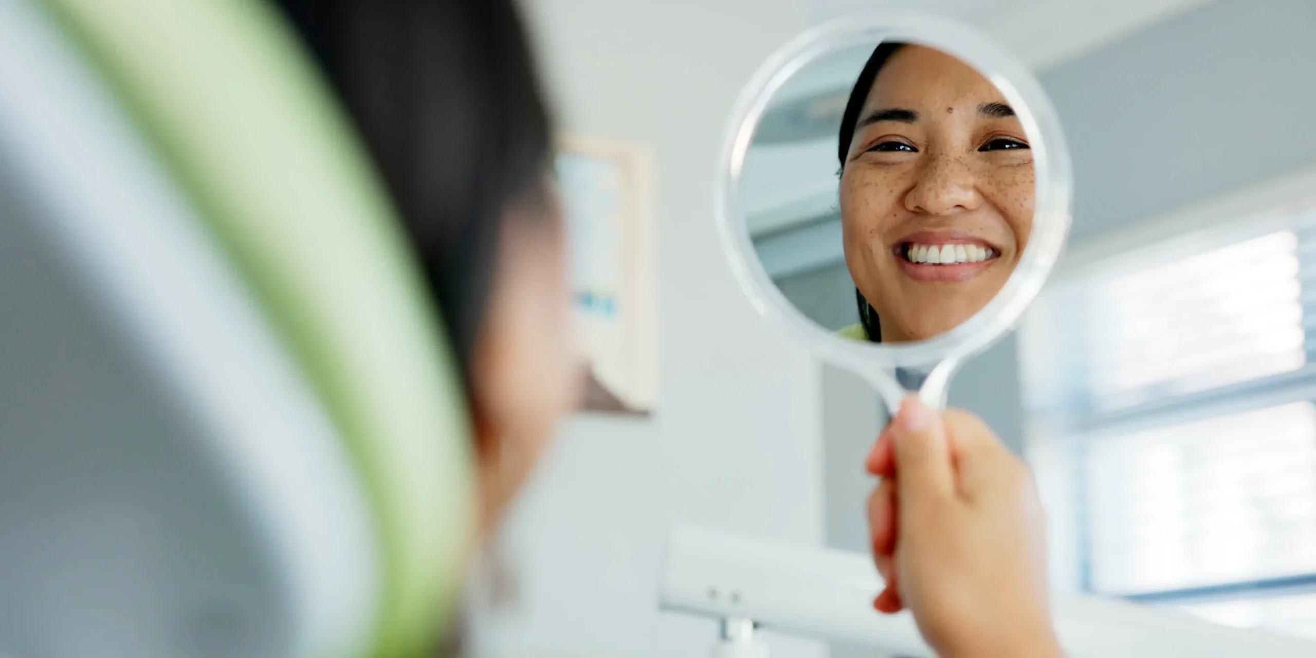 A smiling woman checks out her teeth on a handheld mirror