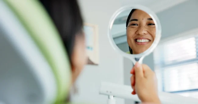 A woman looks at her smile on a mirror A smiling woman checks out her teeth on a handheld mirror