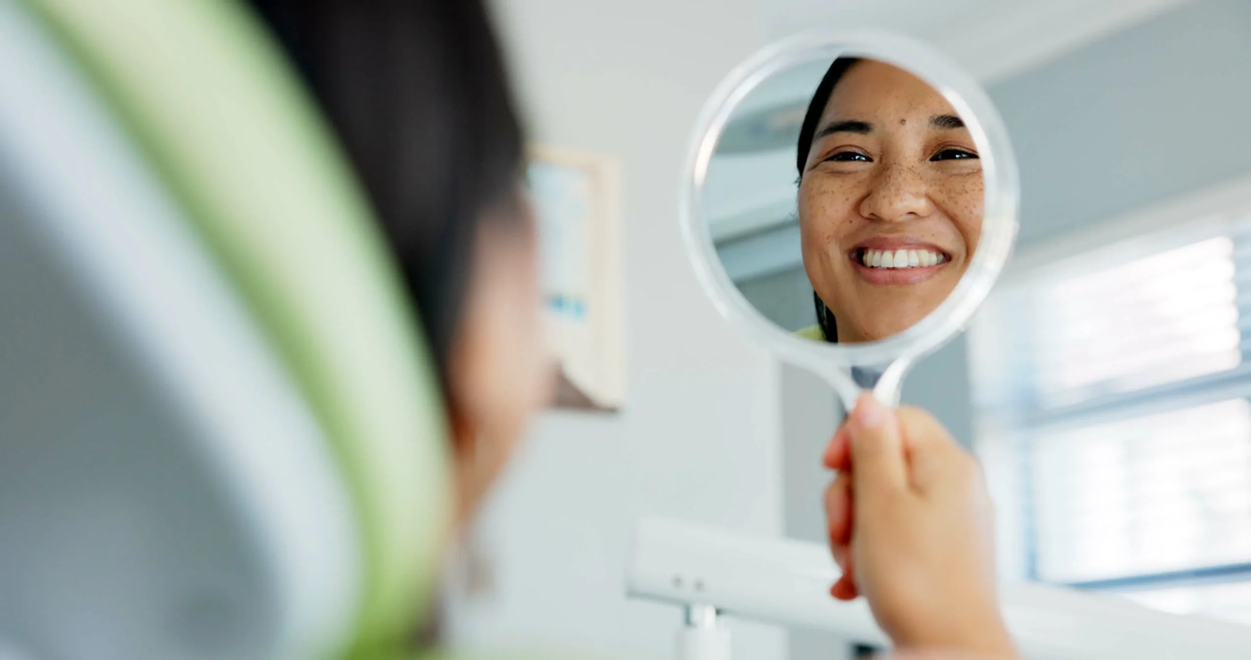 A smiling woman checks out her teeth on a handheld mirror